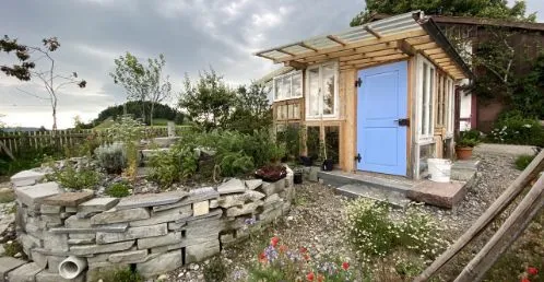 Ein rustikales Gartenhaus mit blauer Tür, umgeben von einer Kräuterschnecke und bunten Blumen