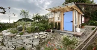 Ein rustikales Gartenhaus mit blauer Tür, umgeben von einer Kräuterschnecke und bunten Blumen